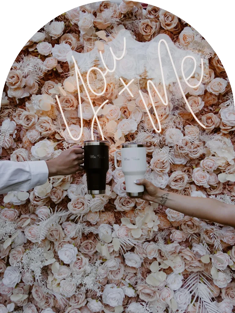 A neon You and Me sign over a blush flower wall, with Mr. and Mrs. mugs raised in a toast
