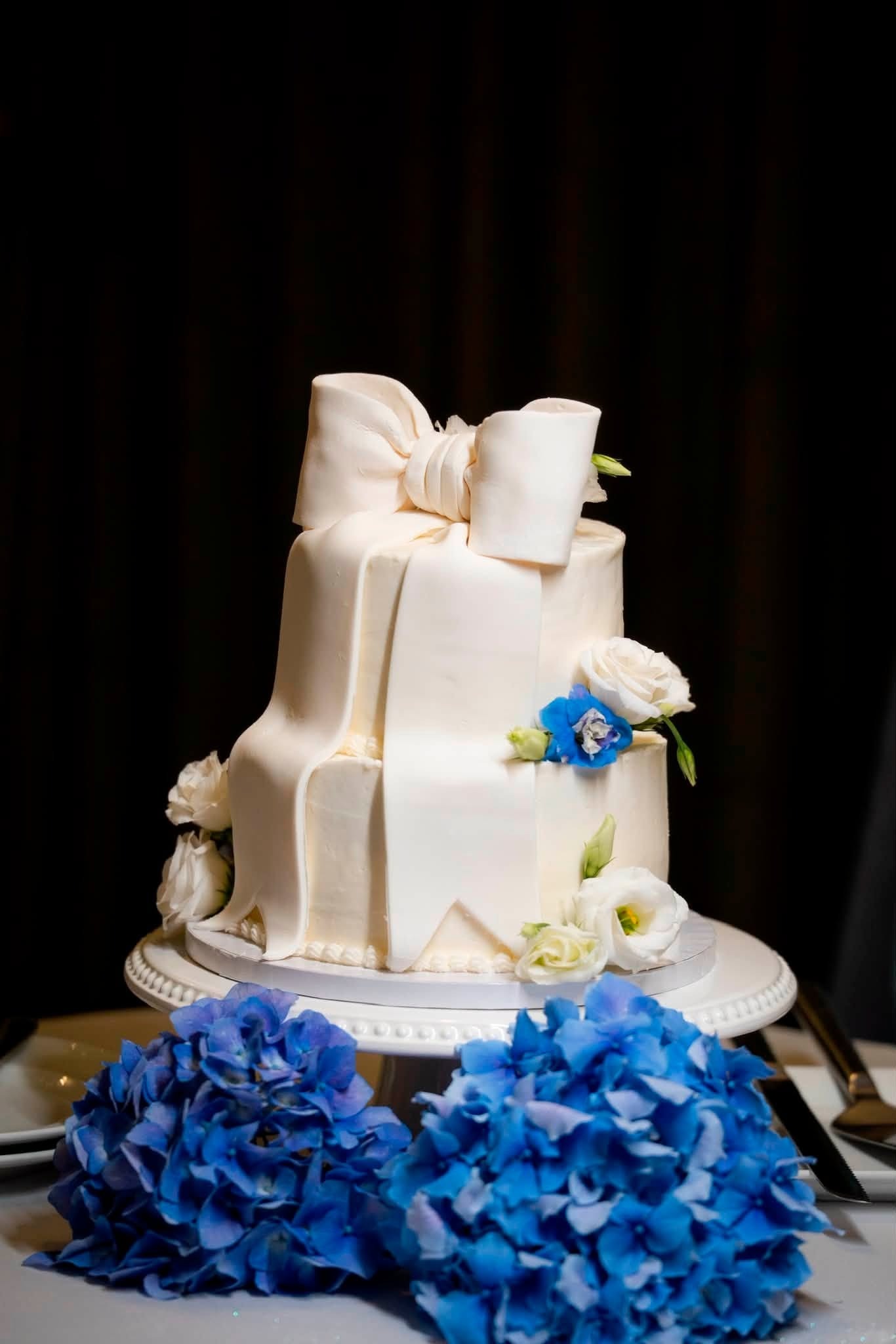 White wedding cake with a large bow on top, surrounded by blue hydrangeas