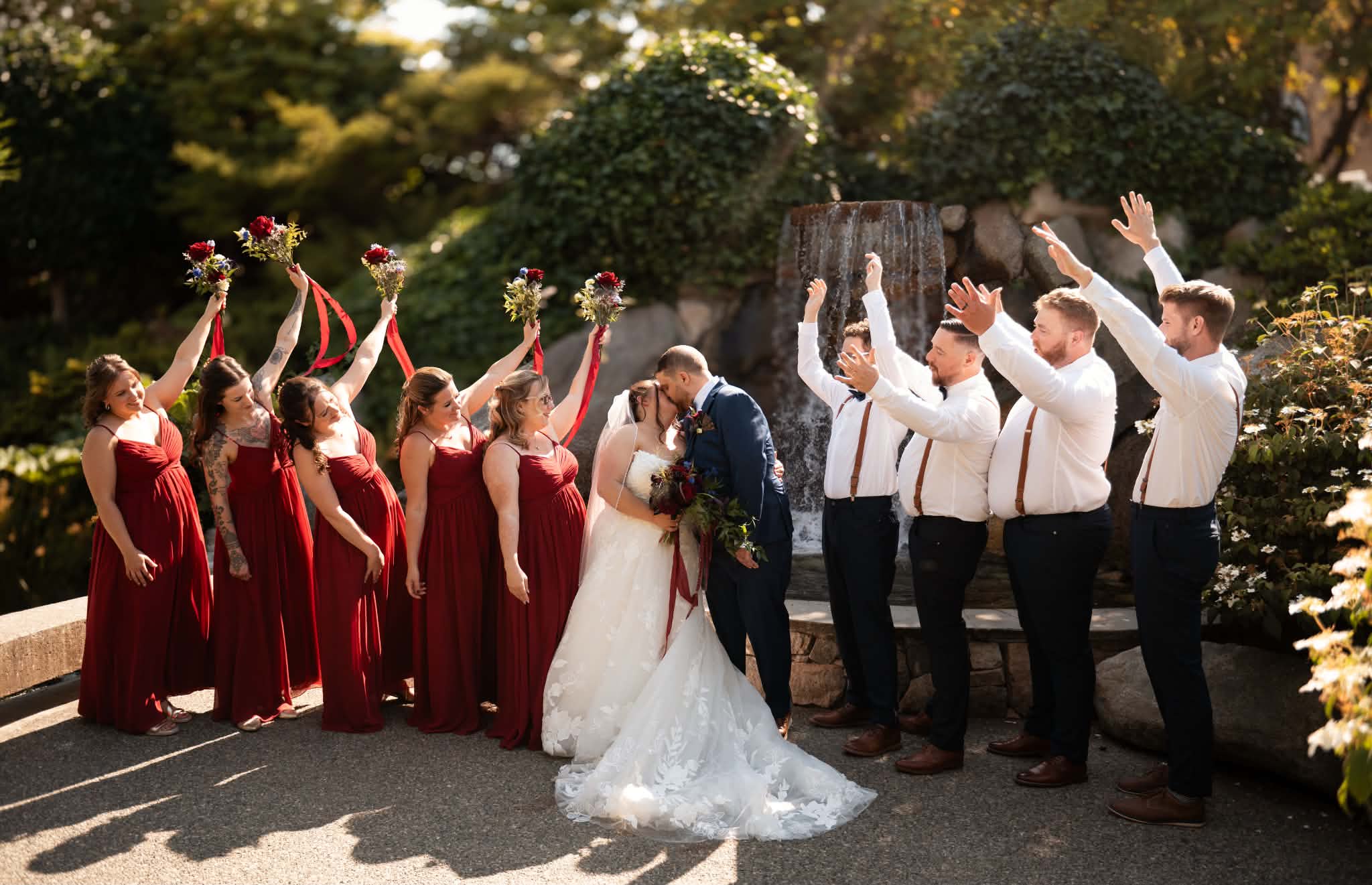 Bridesmaids in red raising their bouquets and groomsmen cheering as Emily and Sheldon kiss by the waterfall