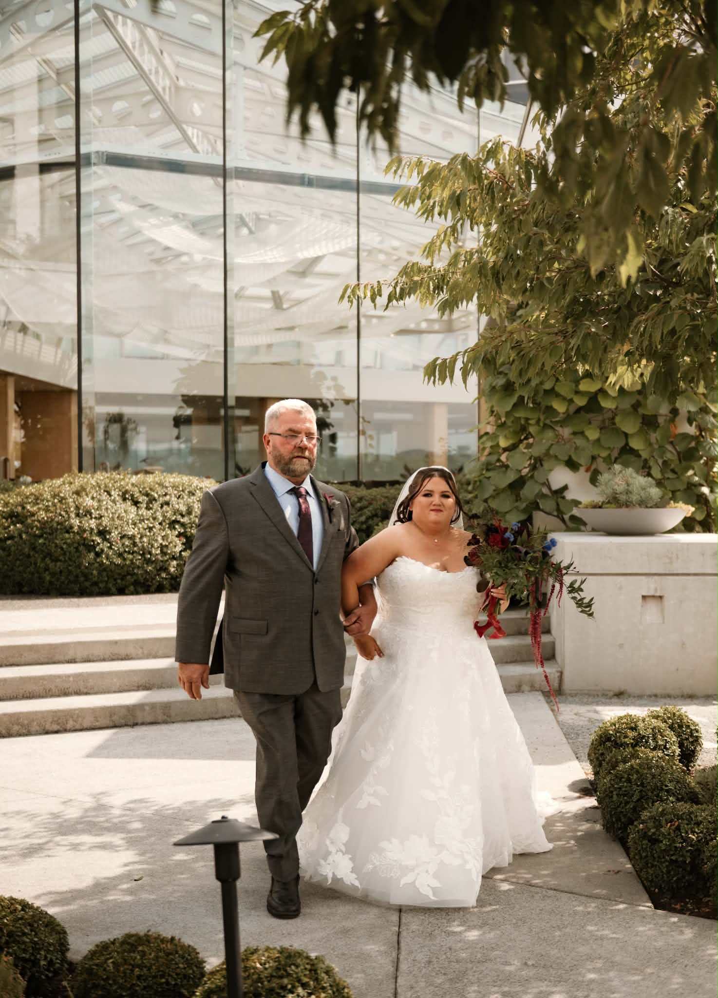 Emily walking down the aisle with her dad in the Laurel Point gardens, holding a burgundy bouquet